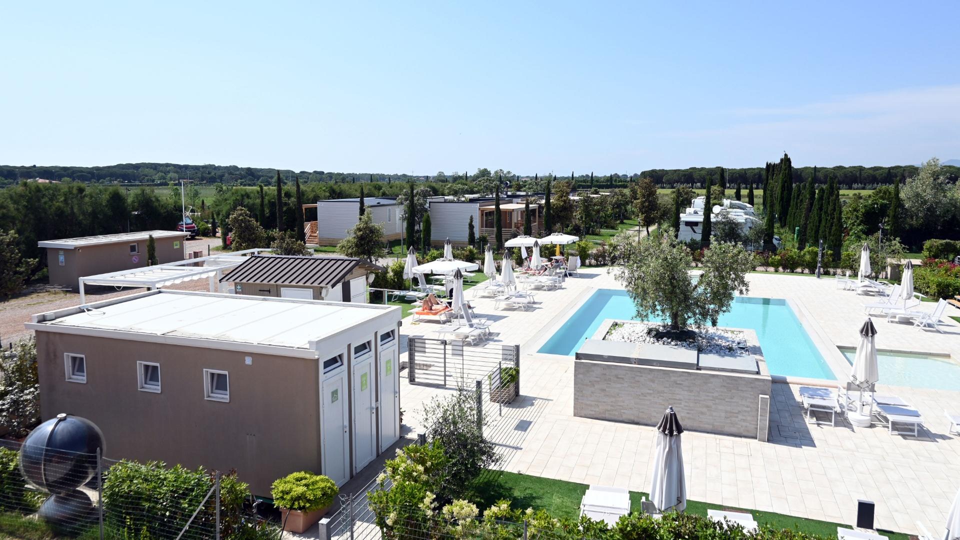 Villaggio turistico con piscina, lettini, ombrelloni e case mobili in un ambiente verde.